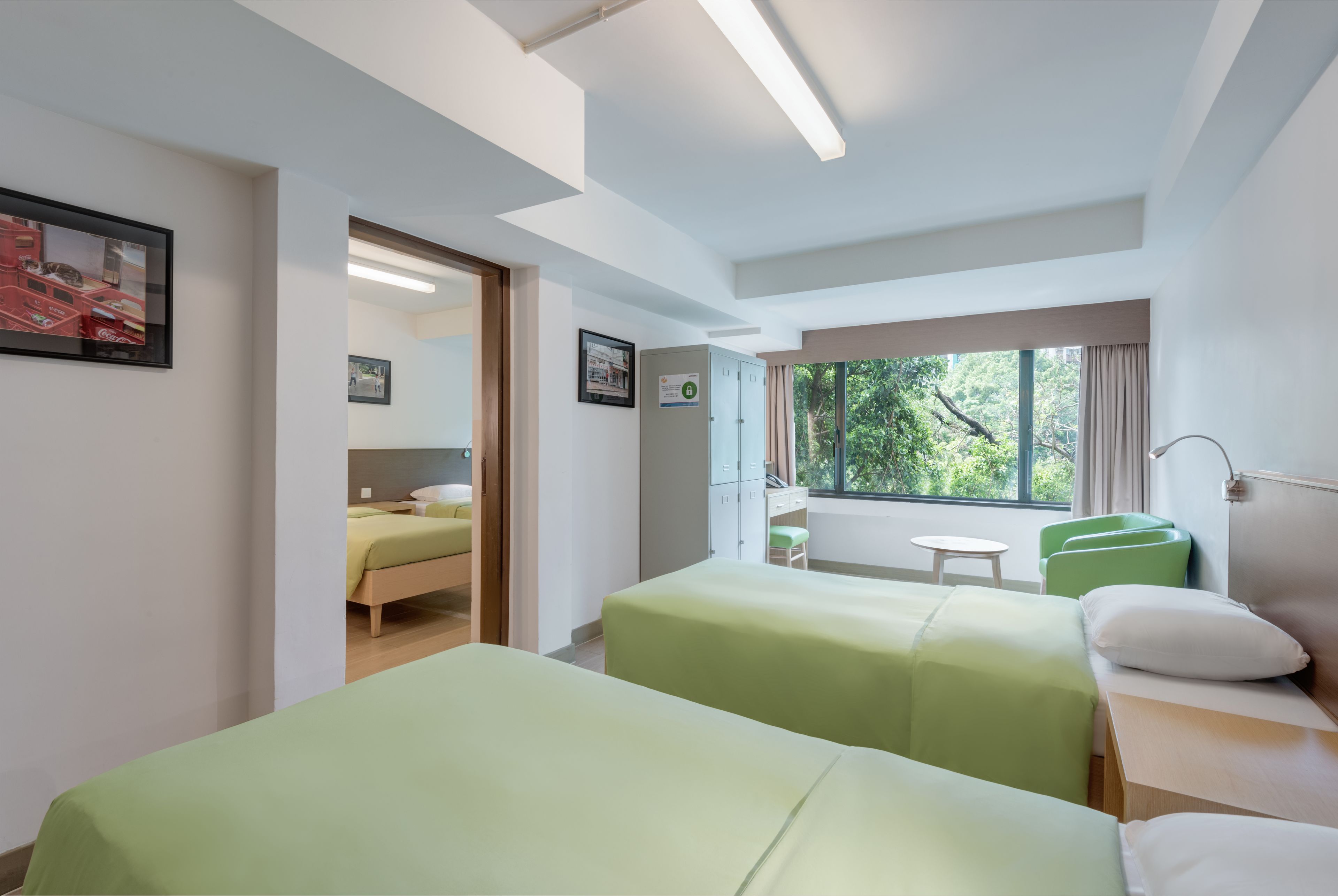 A hotel room with two beds, a small table, a chair, a window with a view of trees, and a cabinet.