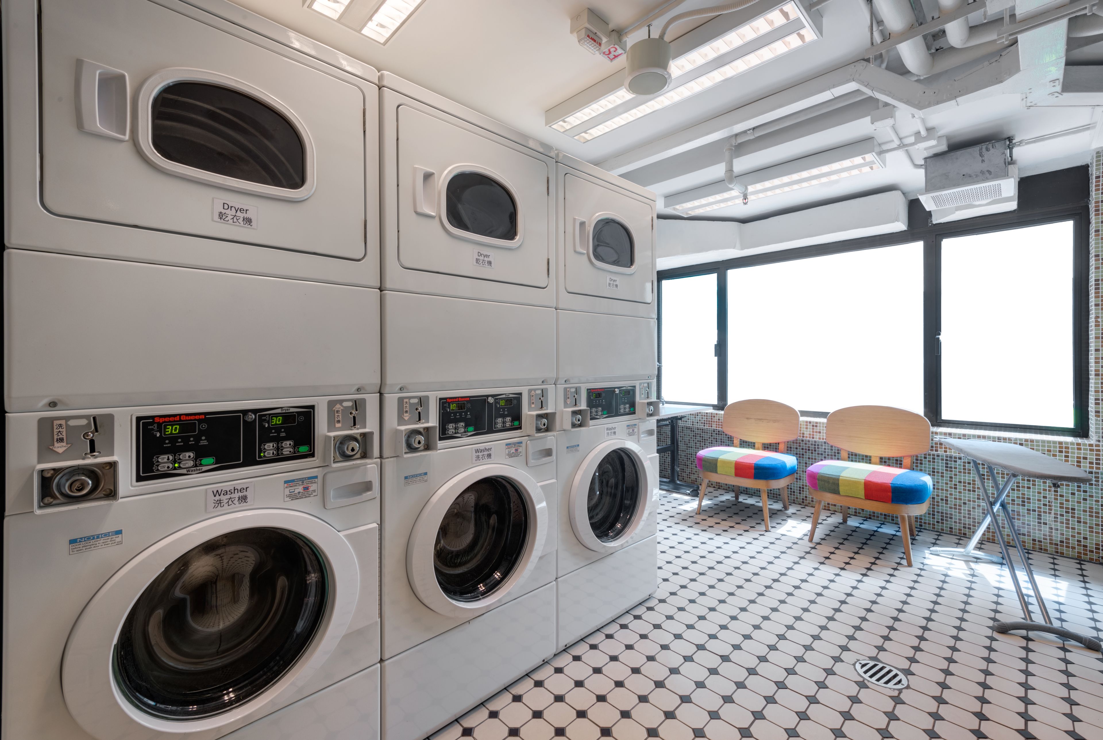 A laundry area with multiple washing machines and dryers, a tiled floor, and two chairs near a window.