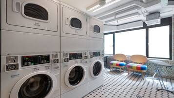 A laundry area with multiple washing machines and dryers, a tiled floor, and two chairs near a window.