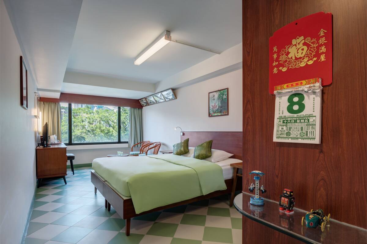 A hotel room with a bed, a desk, a chair, a window, and a calendar on the wall.