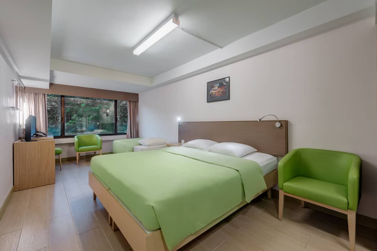 A modern hotel room with a large bed, a green chair, a desk, and a window with curtains.