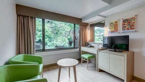 Ein Zimmer mit großem Fenster, Schreibtisch, Stuhl, Fernseher und kleinem Tisch.