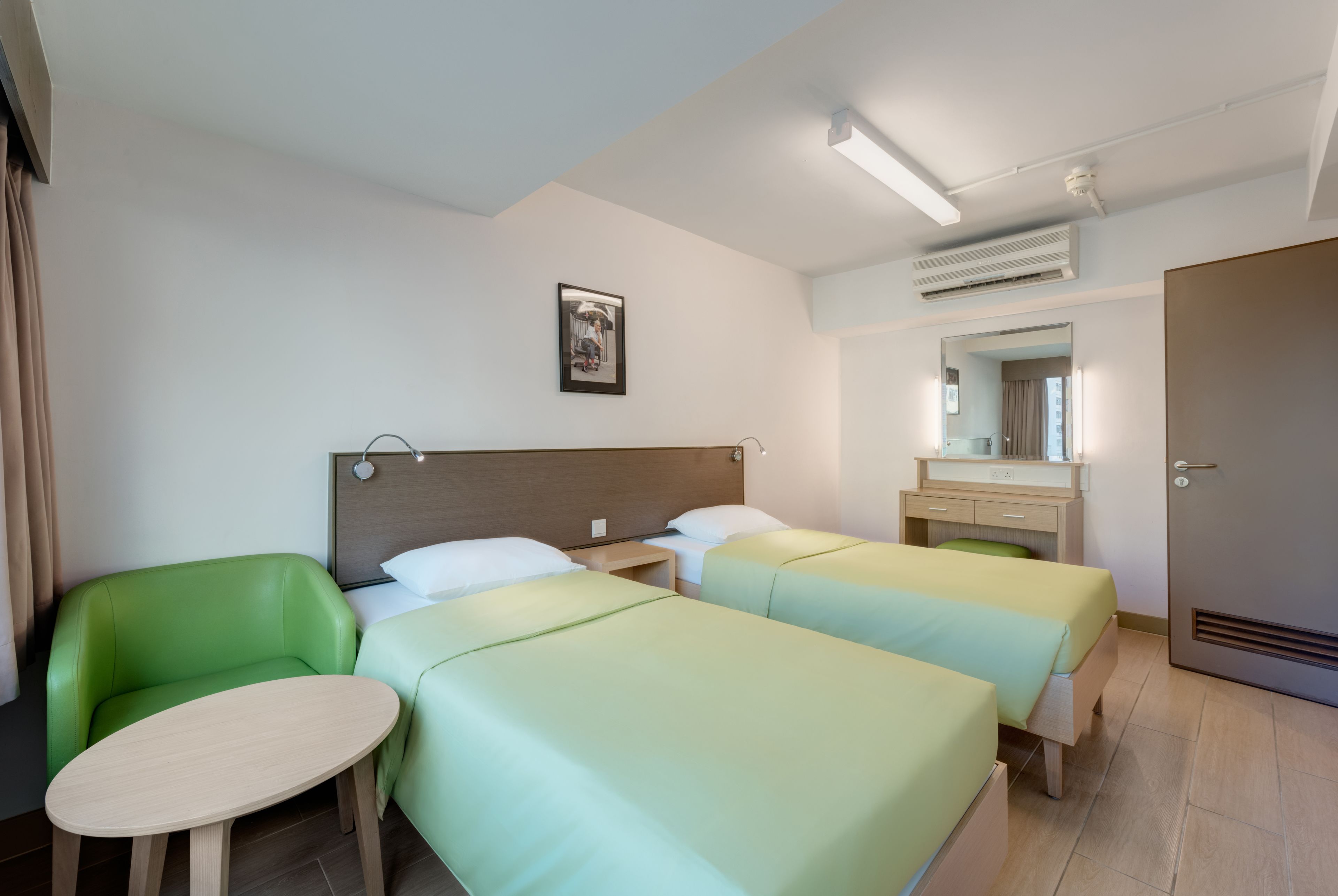 A hotel room with two beds, a green chair, a small table, a bedside table, a mirror, and a door.