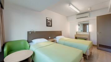 A hotel room with two beds, a green chair, a small table, a bedside table, a mirror, and a door.