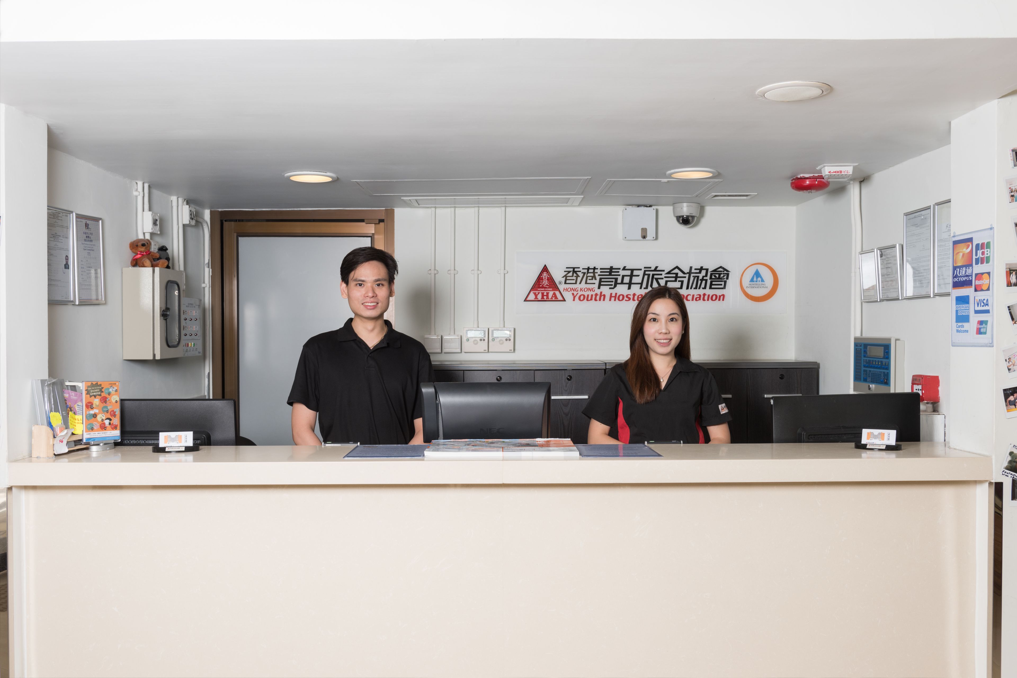 A reception desk with two staff members, a refrigerator, and a wall-mounted sign for the Hong Kong Youth Hostel Association.