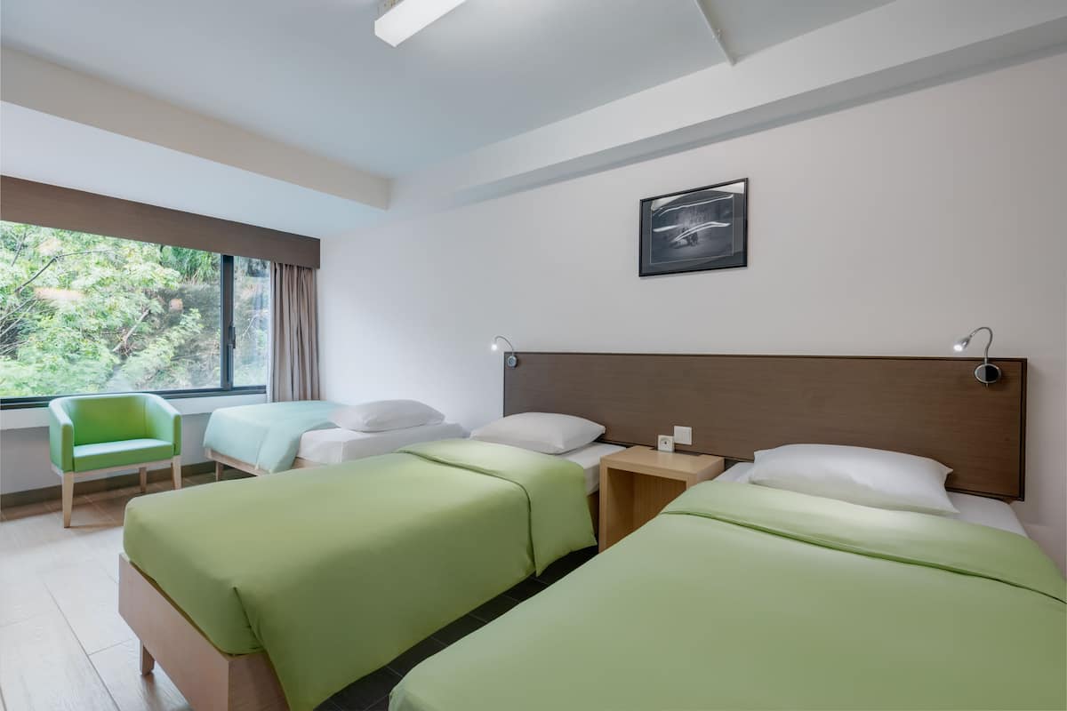 A hotel room with two beds, a green chair, a wooden nightstand, and a window with a view of trees.