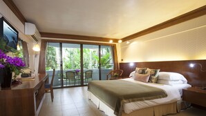 A hotel room with a bed, a desk, a chair, and a balcony with a view of greenery. - Safari Beach Hotel (Patong)