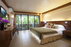 A hotel room with a bed, a desk, a chair, and a balcony with a view of greenery.