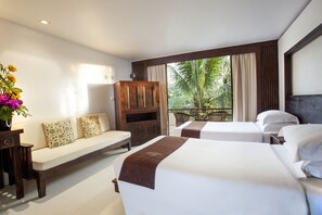 A hotel room with a sofa, two beds, a wooden cabinet, and a balcony with a view of palm trees. - Safari Beach Hotel (Patong)