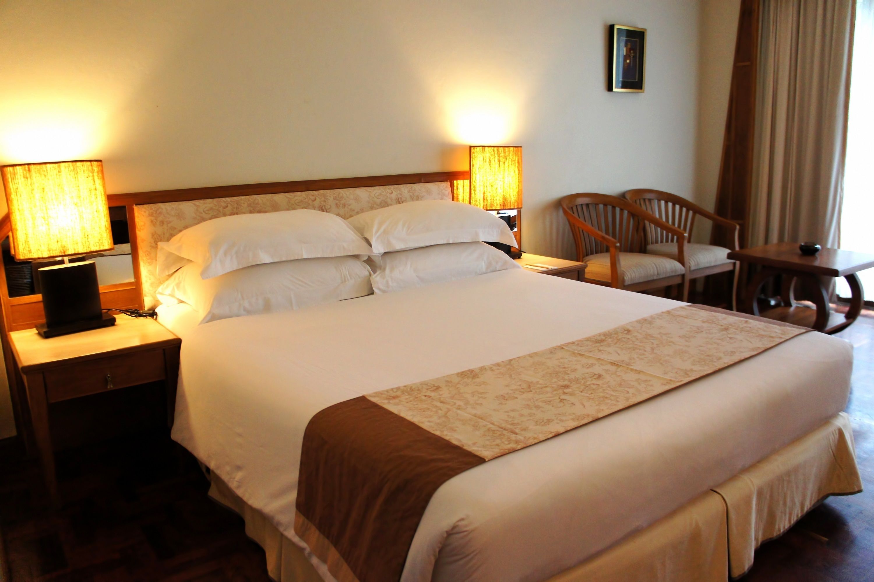 A neatly made bed with a brown accent throw, two bedside lamps, a wooden nightstand, and a chair by the window.