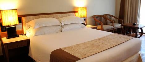 A neatly made bed with a brown accent throw, two bedside lamps, a wooden nightstand, and a chair by the window.