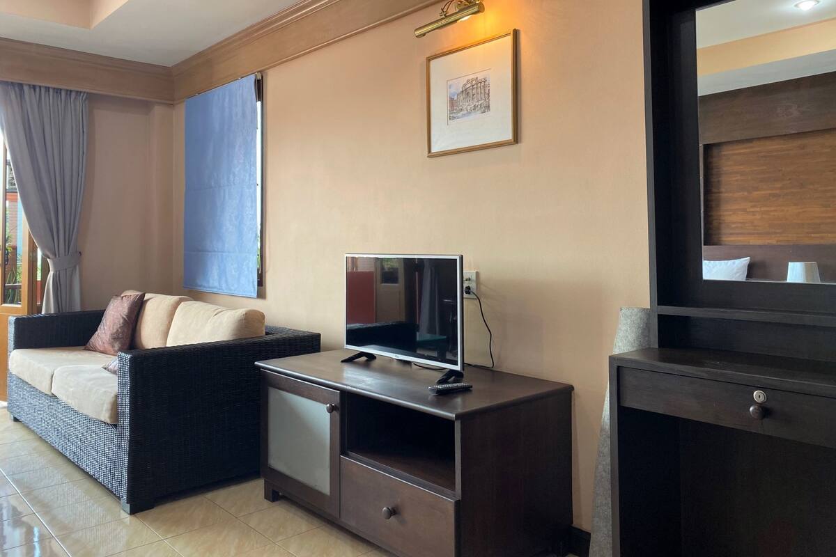 A hotel room with a sofa, a small TV on a wooden cabinet, a framed picture on the wall, and a large mirror.