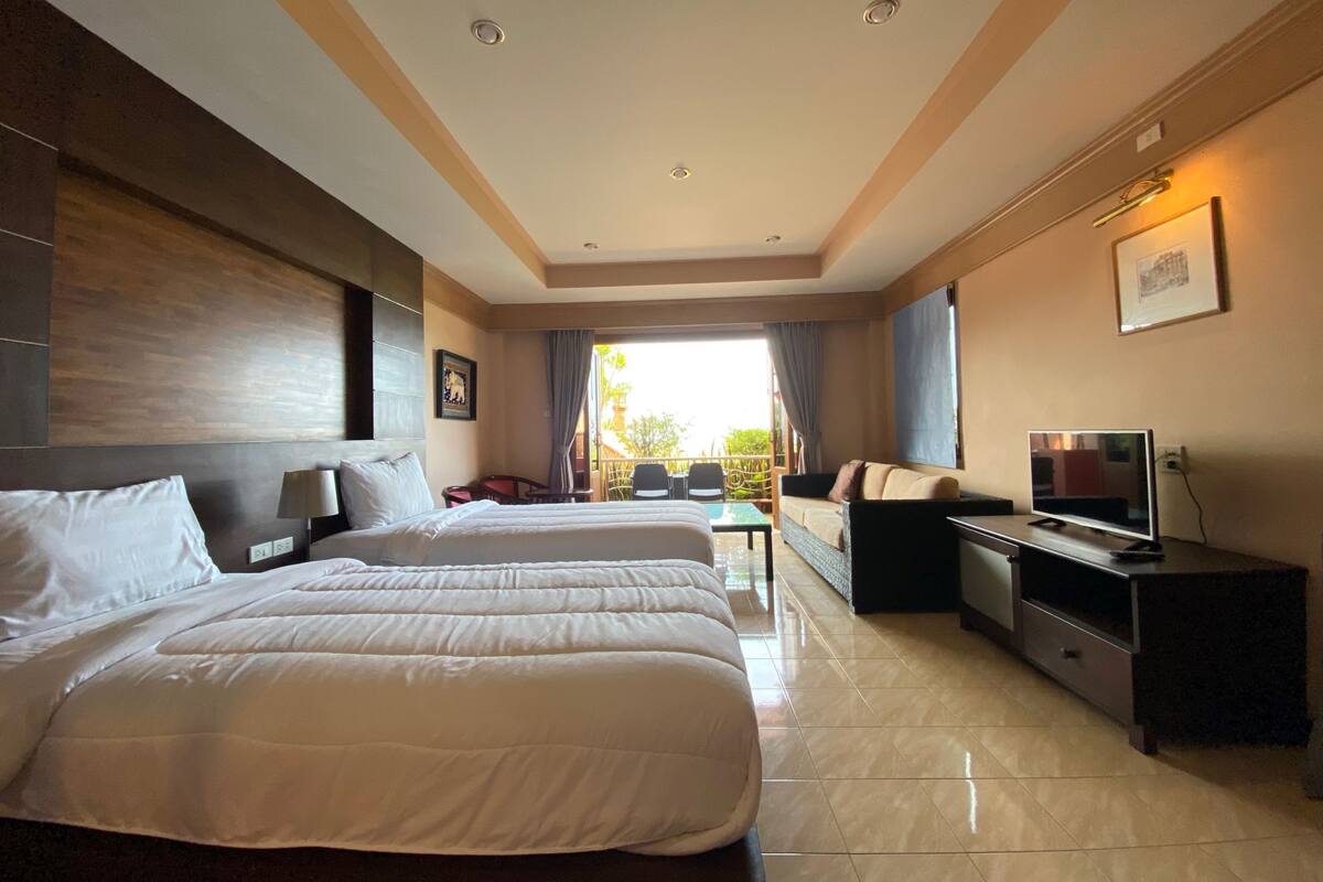 A hotel room with two beds, a TV, a desk, and a balcony with outdoor seating.