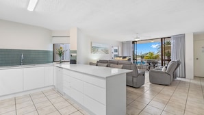 A modern kitchen with a central island, a dining area with a sofa and chairs, and a balcony with a view. - Alexandria Apartments (Alexandra Headland)