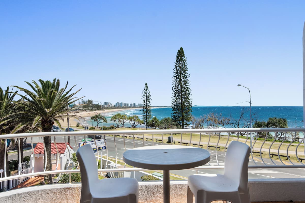 A balcony with two white chairs and a small table, overlooking a beach and ocean with palm trees and a clear sky.