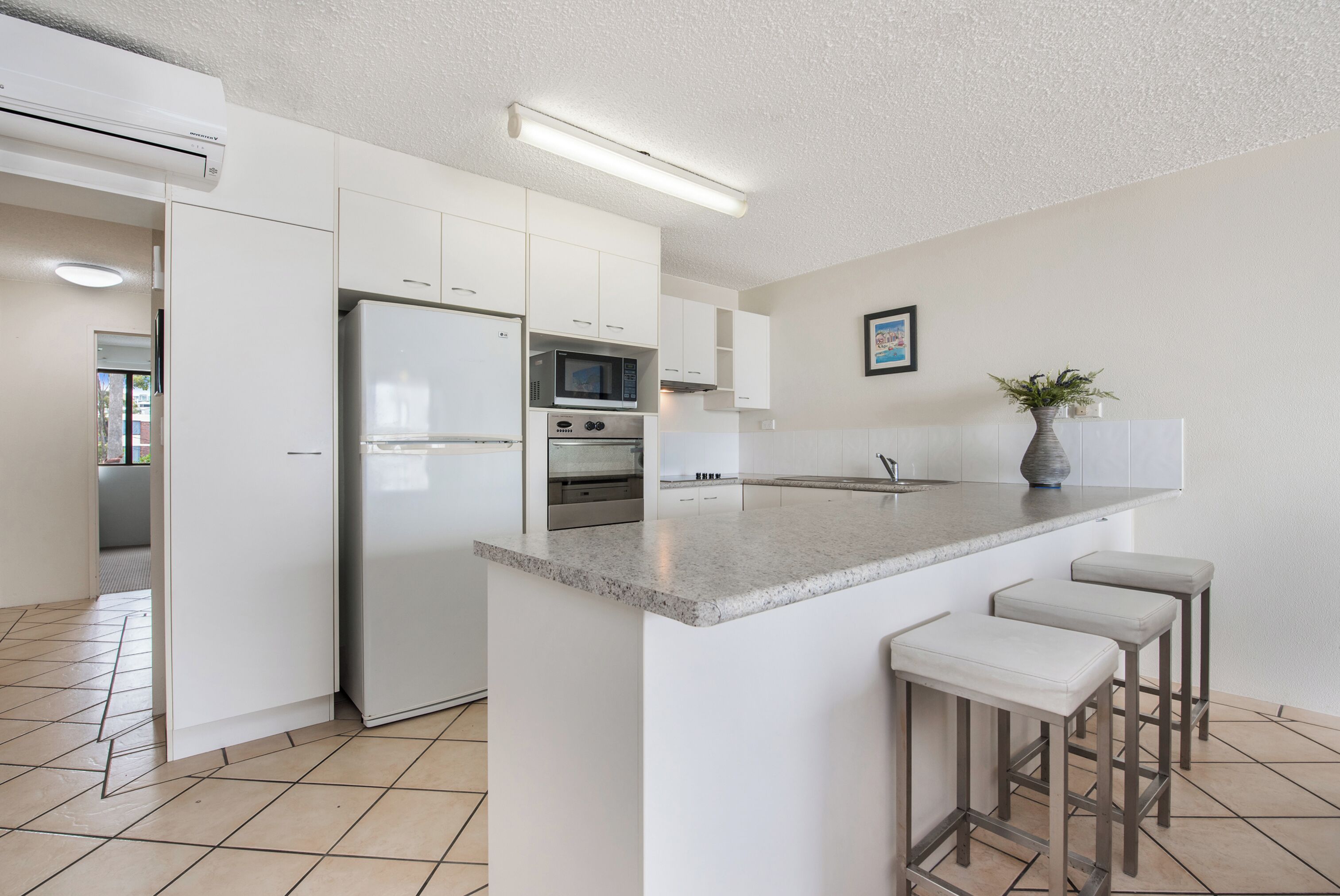 A modern kitchen with white cabinets, a stainless steel refrigerator, an oven, a microwave, and a granite countertop. There are three bar stools and a potted plant on the countertop.