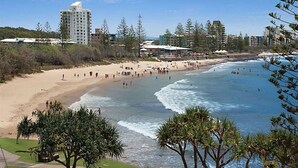 A beach with a hotel in the background, a sandy shore, and trees in the foreground. - Alexandria Apartments (Alexandra Headland)