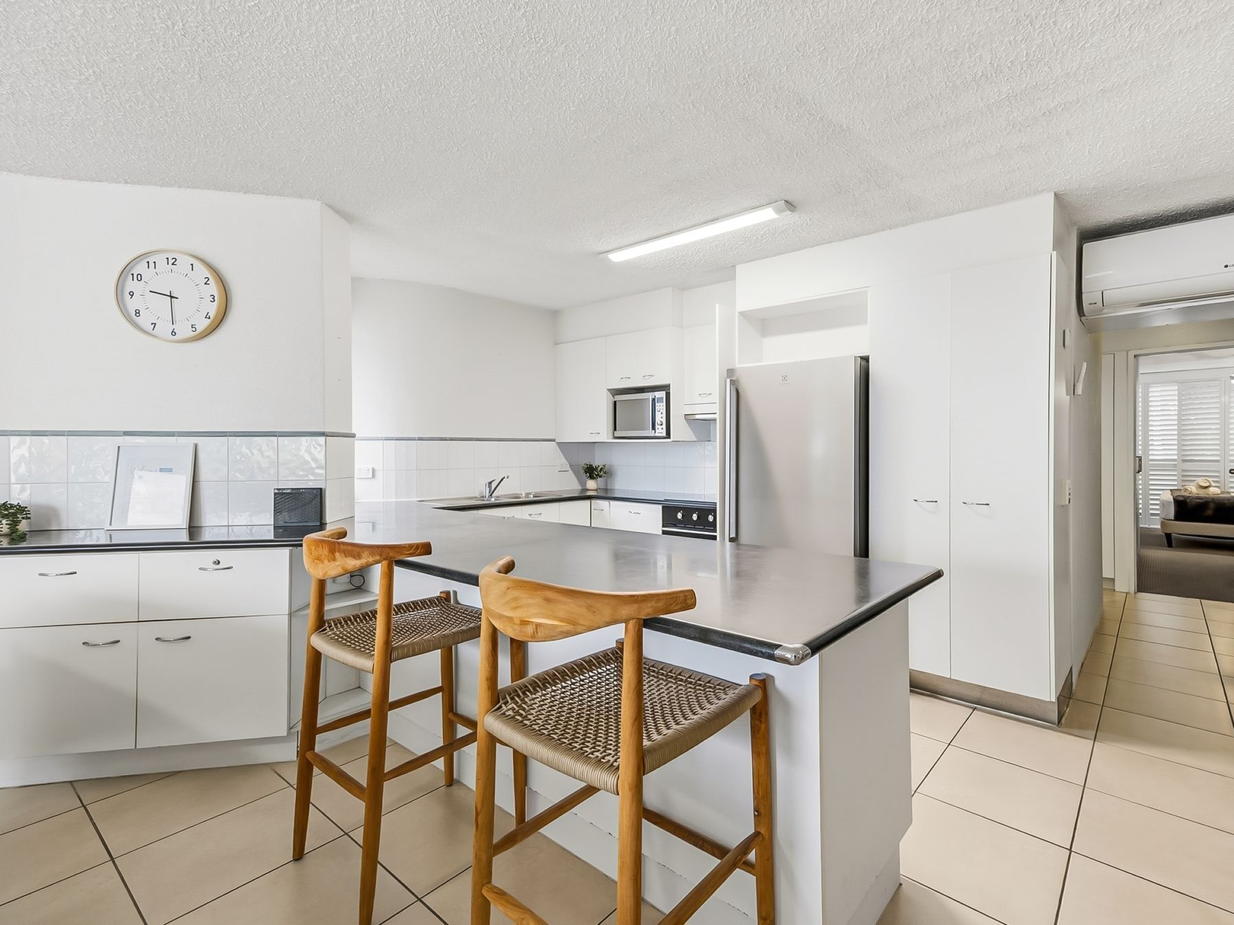 Une cuisine moderne avec des meubles blancs, un îlot central doté de tabourets, un four à micro-ondes, un réfrigérateur et une horloge murale.
