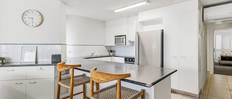 A modern kitchen with white cabinetry, a central island with bar stools, a microwave, a refrigerator, and a clock on the wall.