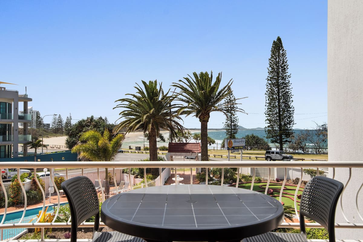 A balcony with a round table and two chairs, overlooking a pool and a beach.
