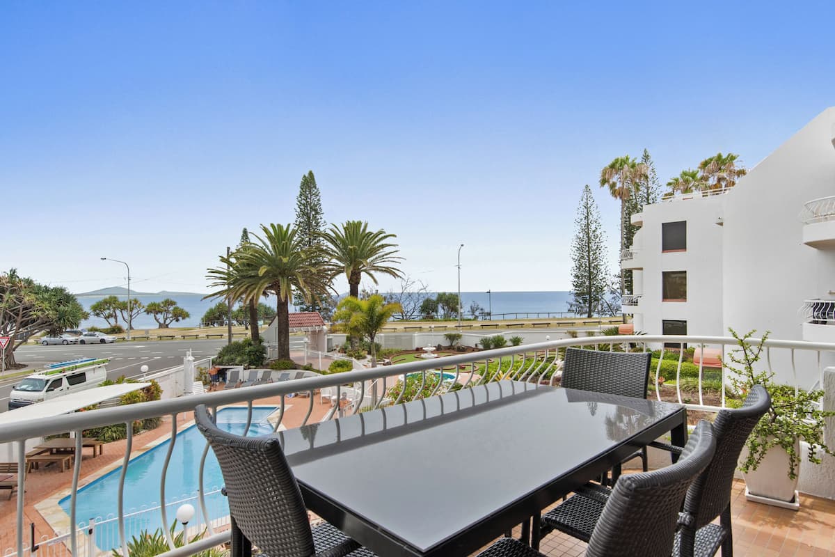 A balcony with a pool, dining table, and chairs, overlooking a coastal area with palm trees and a clear sky.