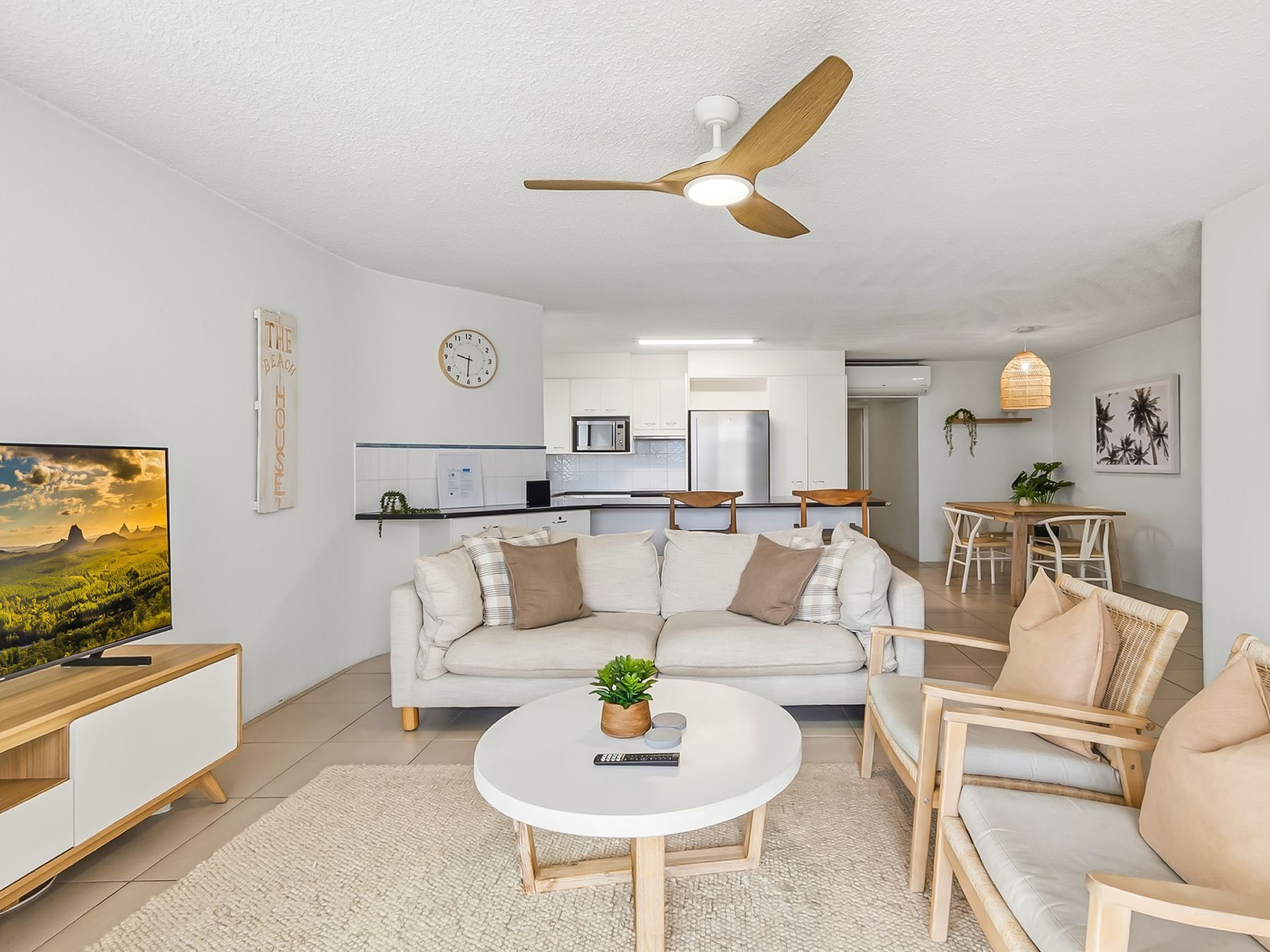A modern living room with a sofa, coffee table, and chairs, a kitchen in the background, and a ceiling fan.