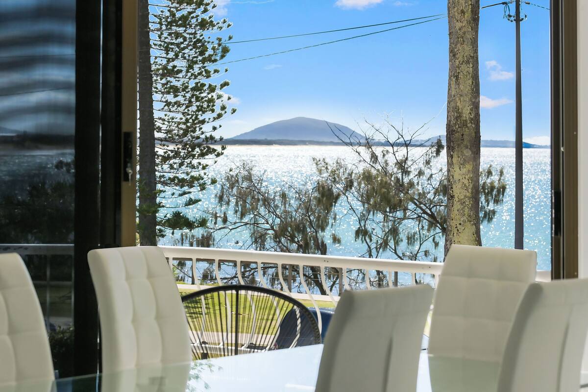 A view from a window with a table and chairs, overlooking a body of water and a distant mountain.