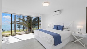 A bedroom with a large bed, a nightstand, a window with a view of palm trees and the sea, and a ceiling light. - Alexandria Apartments (Alexandra Headland)