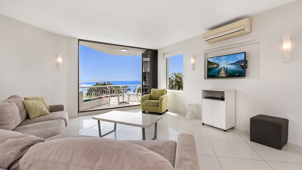 A modern living room with a large window, a flat-screen TV, and a sofa. - Alexandria Apartments (Alexandra Headland)