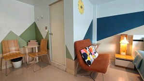 A hotel room with a bed, a nightstand, a lamp, two chairs, a small table with a stack of towels, and a door. - Limburi Hometel (Patong)