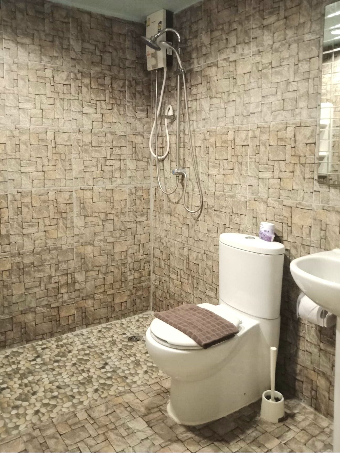 A bathroom with a stone-patterned wall, a toilet with a brown mat, a shower head, and a sink with a toilet brush.