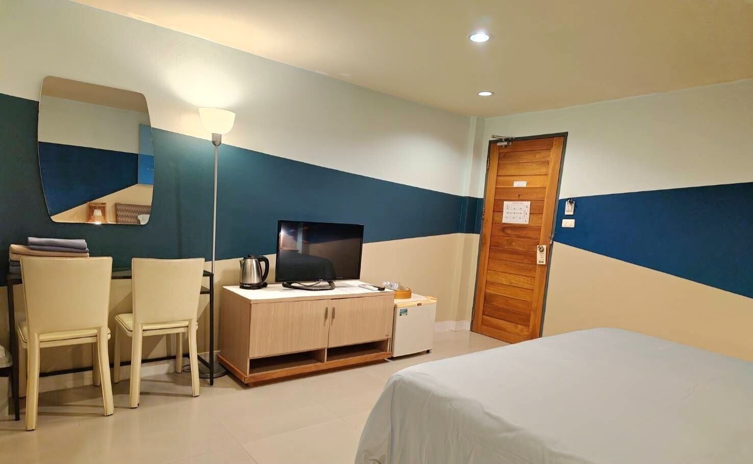 A hotel room with a bed, a desk with a television, a chair, a small refrigerator, and a wooden door.