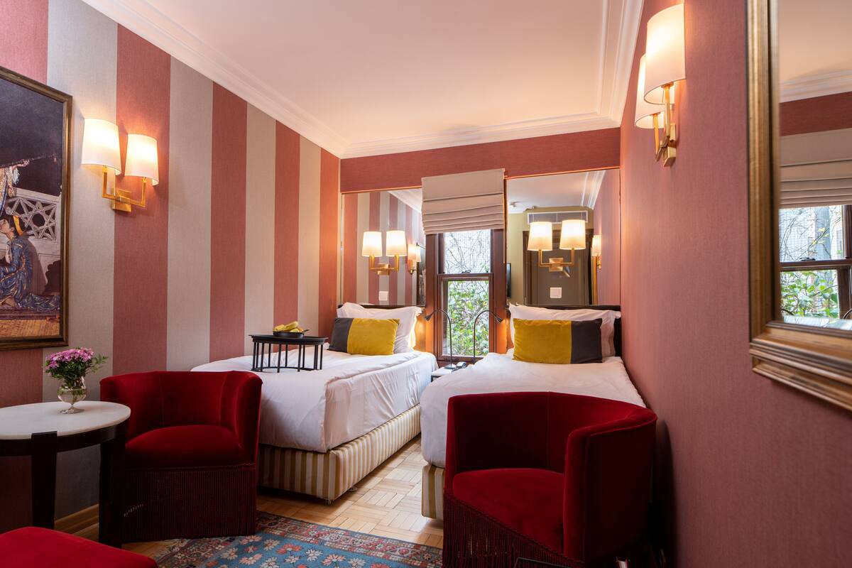 A hotel room with two beds, a small table with a vase of flowers, a red armchair, a red sofa, and a bookshelf.
