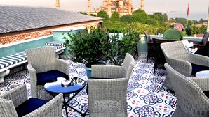 Terraza en la azotea con mobiliario de mimbre y suelo estampado, con vistas a un edificio histórico con múltiples alminares.
