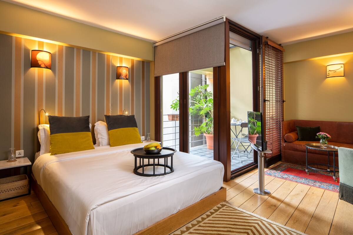 A hotel room with a large bed, bedside tables, a small sofa, a balcony with a potted plant, and a sliding door.