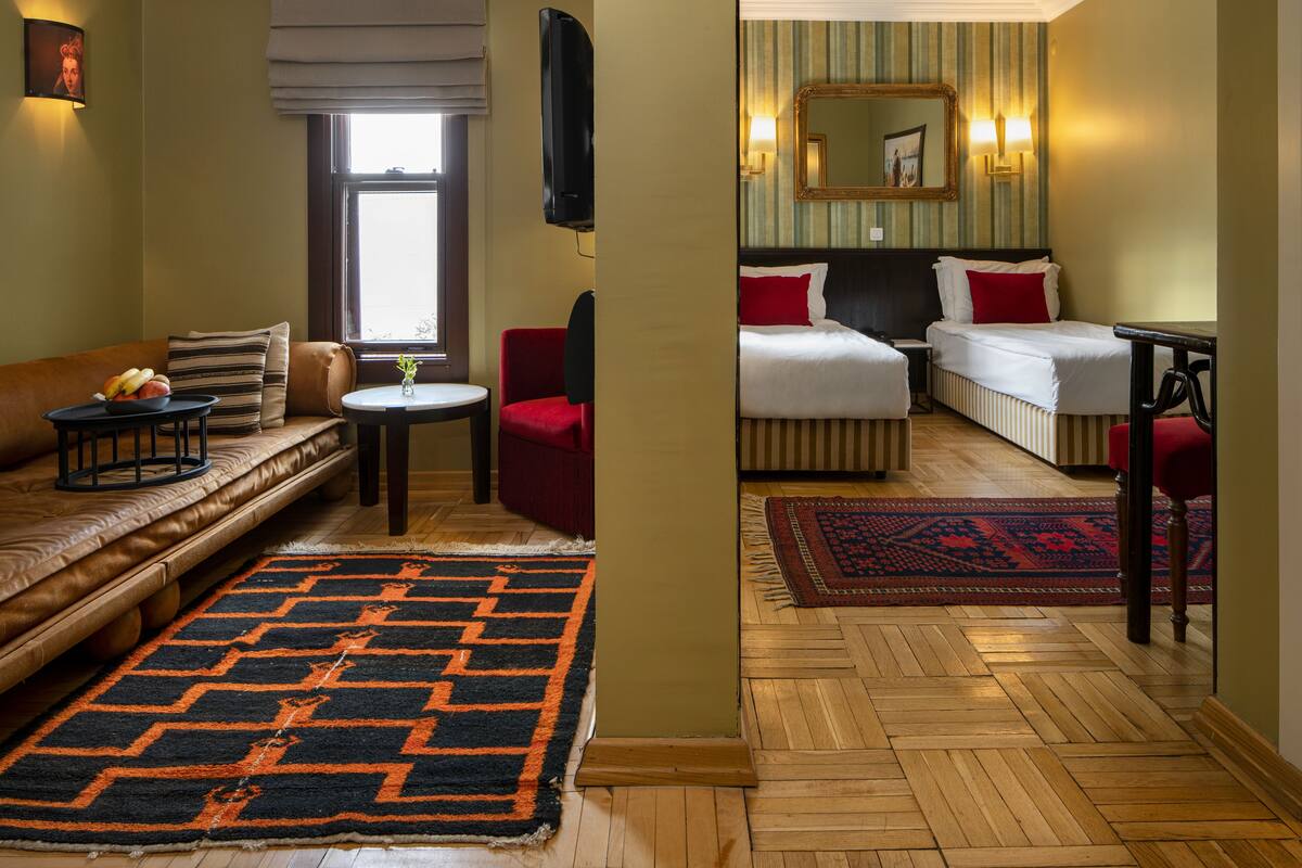 A hotel room with a sofa, a small table with a bowl of fruit, a red chair, two beds with red pillows, a large mirror, and a patterned rug.