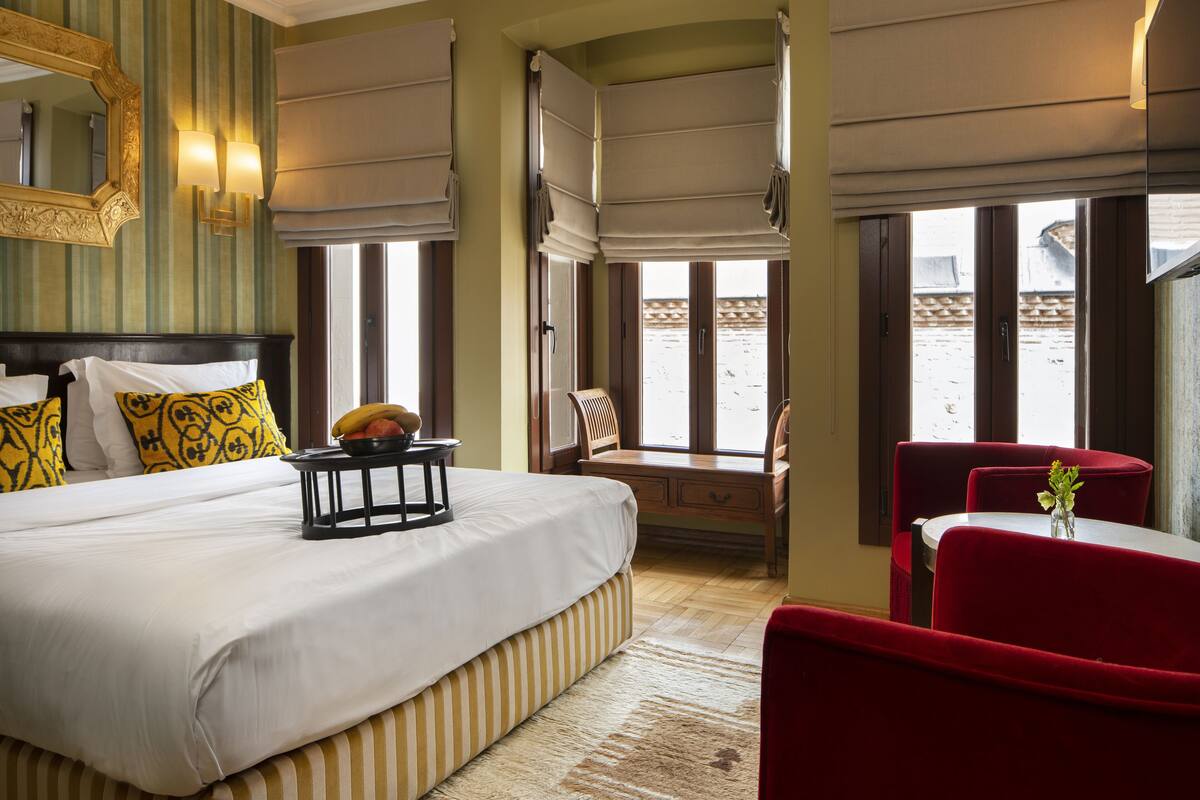 A hotel room with a large bed, a small table with fruit, a red chair, and a view of the beach through the windows.