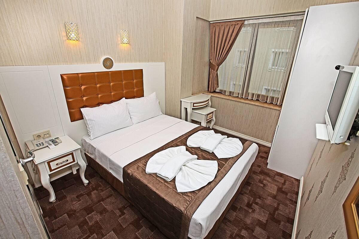 A hotel room with a large bed, a bedside table with a phone, a small table with a lamp, a window with curtains, and a television on a stand.