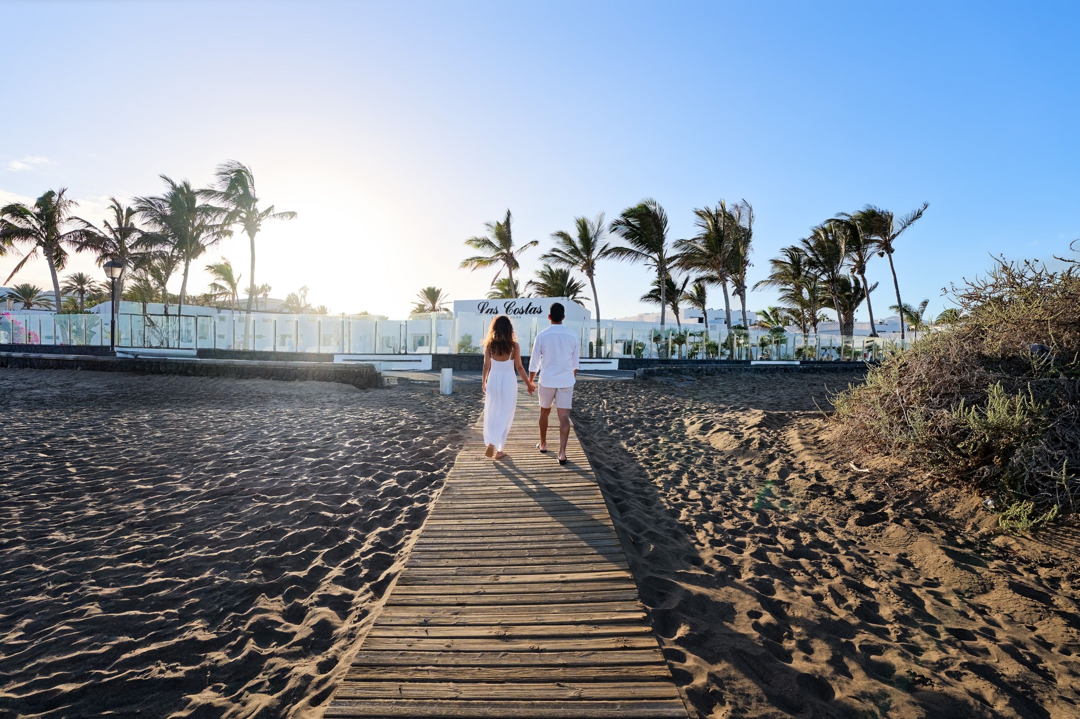 A wooden boardwalk leading to a beach area with palm trees and a sign reading 'Pans Costas.'