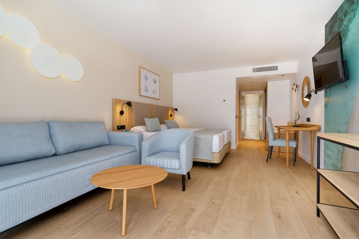 A modern hotel room with a sofa, a small table, a bed, a TV, and a wall-mounted artwork.