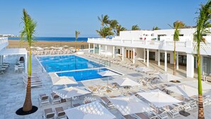 A resort pool area with sun loungers and umbrellas, a clear view of the beach, and a building with a white facade.