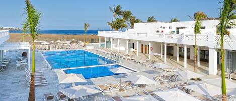 Zona de piscina en un resort con tumbonas y sombrillas, vista despejada a la playa y un edificio de fachada blanca.