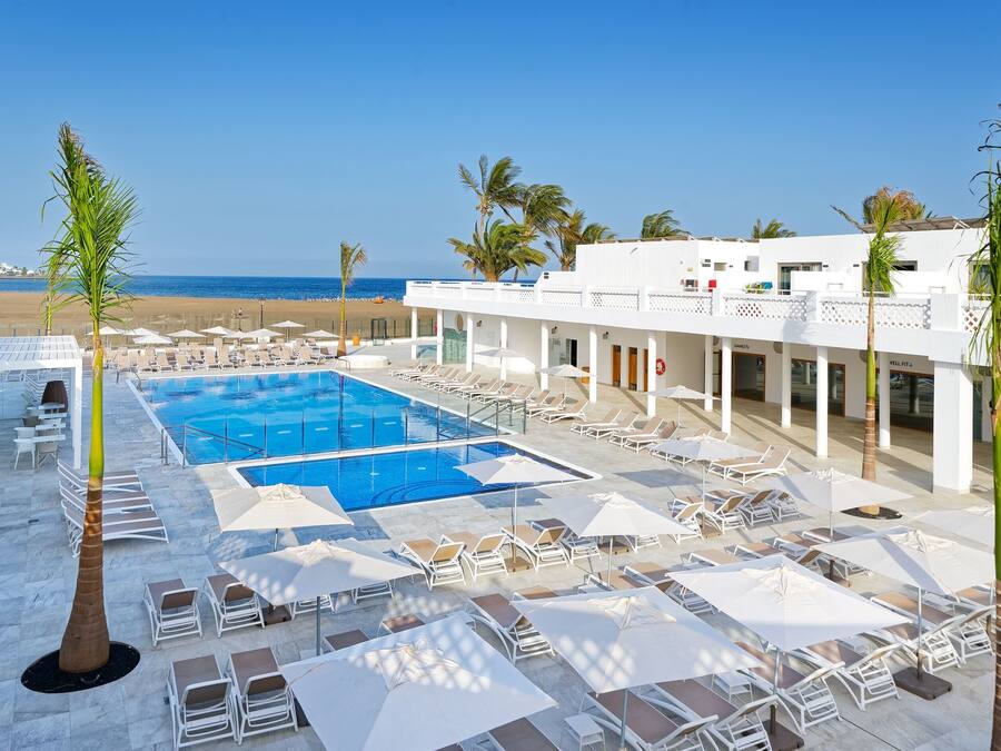 A resort pool area with sun loungers and umbrellas, a clear view of the beach, and a building with a white facade.