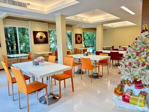 A modern dining area with white tables and orange chairs, a Christmas tree with gifts, and large windows with a view of greenery. - Calypzo Bangkok (Bangkok)