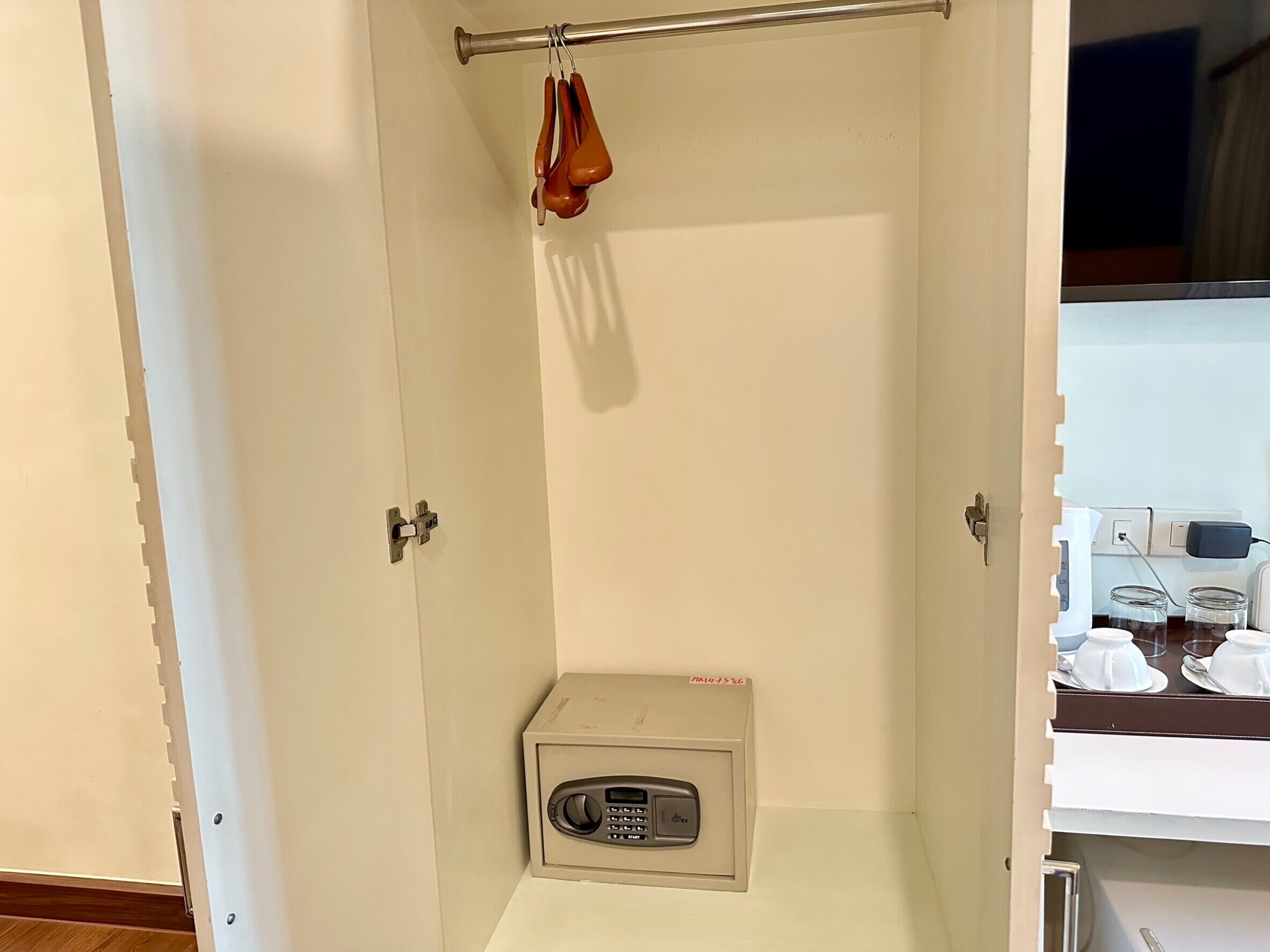 A small, enclosed space with a metal rod and a hanging object, a small metal box with a keypad, and a white shelf with cups and a knife.