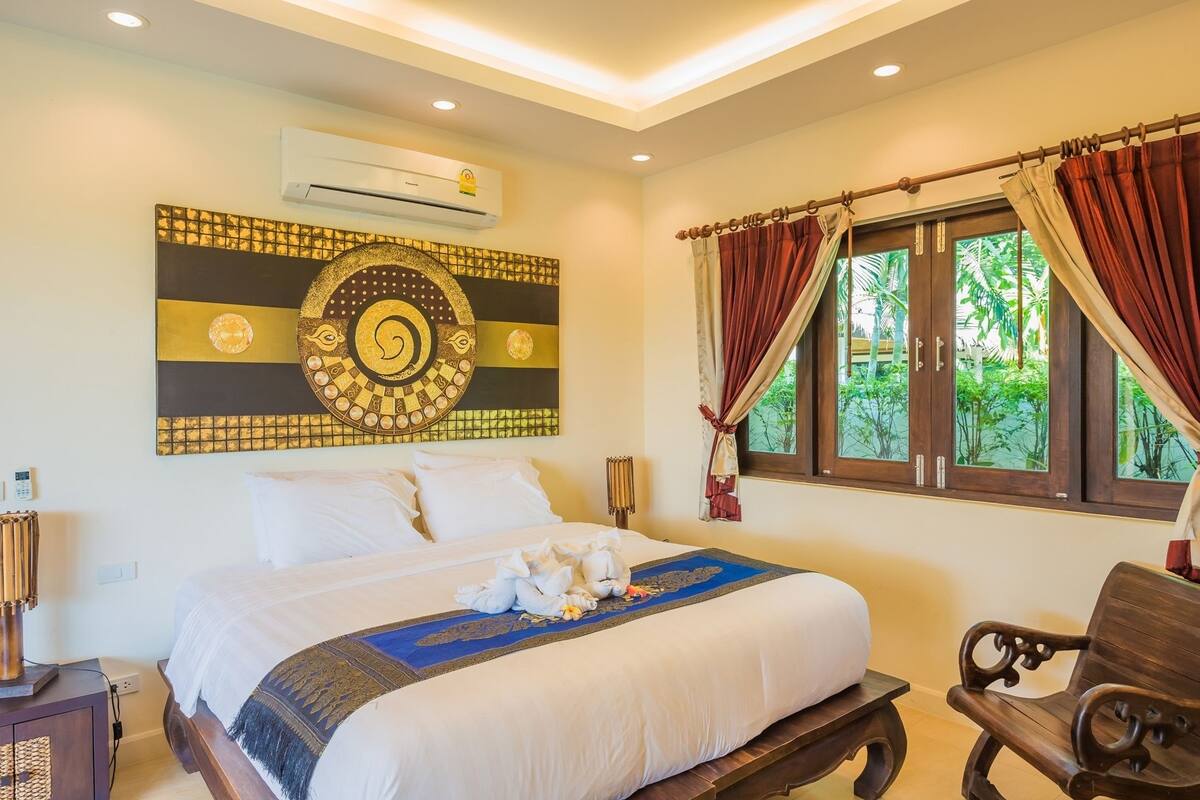 Une chambre avec un grand lit, un élément décoratif mural, un climatiseur, une fenêtre avec des rideaux et un banc en bois.
