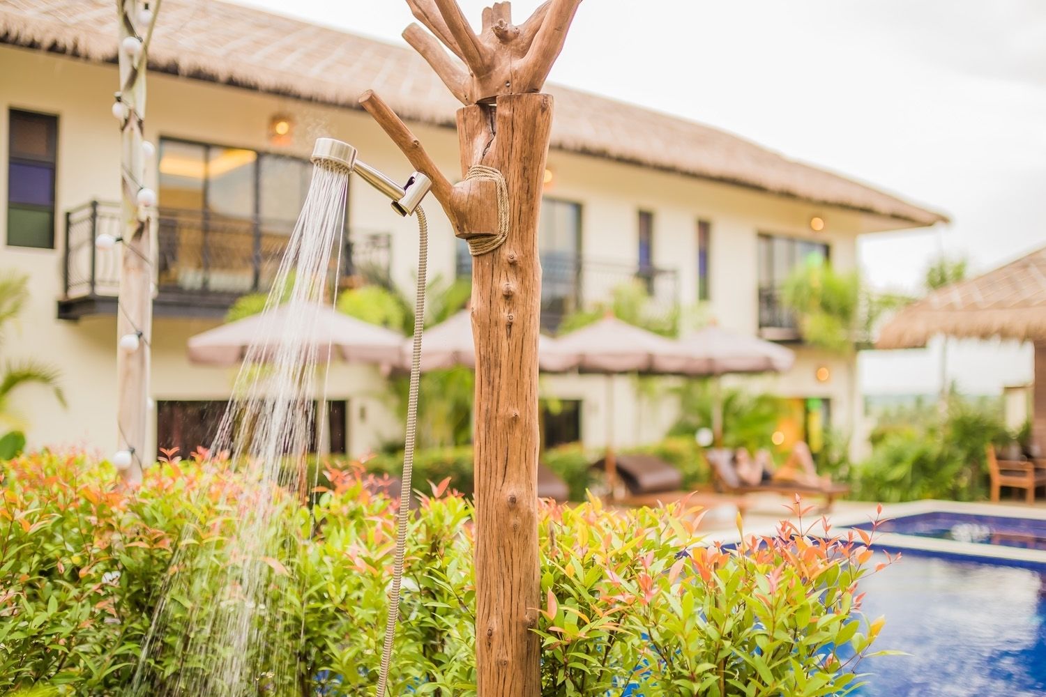 A wooden water feature with a shower head, surrounded by lush greenery and a pool, with a building in the background.