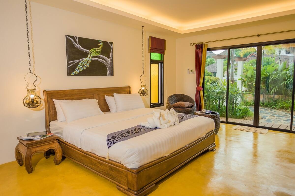 Une chambre spacieuse avec un grand lit, une tête de lit en bois et une vue sur un jardin luxuriant, accessible par une porte-fenêtre coulissante.