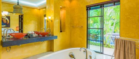 A modern bathroom with a large bathtub, dual sinks with red bowls, a mirror, and a view of greenery through a glass door.
