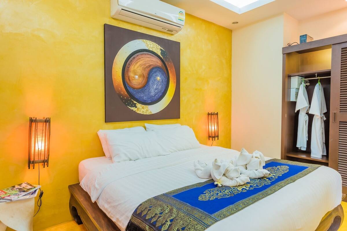 Une chambre d’hôtel avec un lit, un tableau représentant un mandala, une moquette bleue et dorée, et une armoire remplie de vêtements.